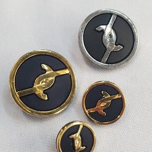 Puede incluir: Cuatro botones ovalados con centro negro y borde dorado o plateado. Cada botón presenta un diseño estilizado dorado o plateado. Los botones varían en tamaño y están dispuestos sobre una superficie blanca.