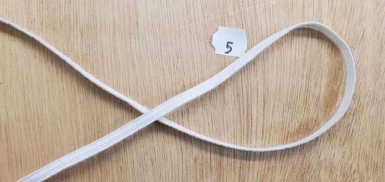 K&ouml;nnte beinhalten: Eine d&uuml;nne, wei&szlig;e, flache elastische Kordel, die in einer Schlaufe angeordnet ist und sich selbst kreuzt. Ein kleines wei&szlig;es Papieretikett mit der in blauer Tinte geschriebenen Zahl 5 befindet sich in der Mitte der Schlaufe als Referenz.