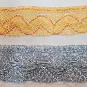 May include: Close-up of two decorative lace trims. The top trim is a light yellow with a scalloped edge and a zig-zag pattern. The bottom trim is a light blue-grey with a similar zig-zag pattern. Both trims are on a white background.