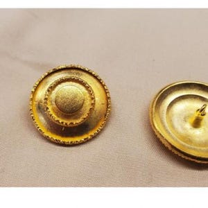 May include: Two gold-toned metal buttons with a raised, circular design. The button on the left has a concentric circle design, while the button on the right has a smooth surface.