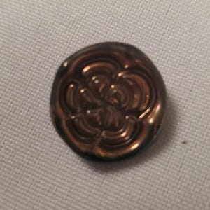 Könnte beinhalten: Ein runder Knopf mit metallisch bronzefarbener Oberfläche und einem erhabenen, geprägten Blumenmuster. Das Design besteht aus konzentrischen, geschwungenen Linien, die eine stilisierte Blütenform bilden und für eine reflektierende Optik sorgen.