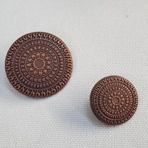 Op de afbeelding: Twee koperkleurige knopen met een gedetailleerd cirkelvormig ontwerp. De grotere knoop heeft een diameter van ongeveer 3,2 cm, de kleinere ongeveer 1,9 cm. De knopen zijn geplaatst tegen een witte stoffen achtergrond.