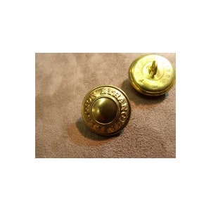 Può includere: Due bottoni color oro con un design circolare in rilievo e testo. I bottoni hanno un anello sul retro per cucire. I bottoni sono su una superficie marrone. Il testo sui bottoni recita "LONDON TERRANCE".