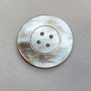 May include: A large, round button with an iridescent sheen. The button features a central, slightly sunken area with four small holes for sewing. The button is set against a neutral, textured background.
