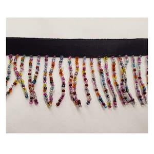 Peut inclure: Un ruban noir avec des franges de perles. La frange présente des brins de perles colorées dans les tons de violet, d'orange, de jaune et de bleu. Les perles sont enfilées ensemble, créant une garniture décorative.