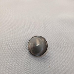 Puede incluir: Un botón redondo gris con un acabado brillante. El botón tiene una forma ligeramente abovedada y una superficie lisa. El botón está sobre un fondo de tela gris claro. Este botón podría usarse para ropa o manualidades.