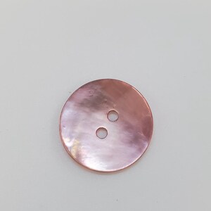 May include: A circular, two-hole button with a smooth, iridescent surface in a soft, dusty rose pink hue. The material displays a subtle, pearlescent sheen that reflects light across its flat, round face, highlighting the natural texture.