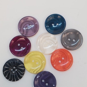 Könnte beinhalten: Eine Sammlung von neun runden Knöpfen in verschiedenen Farben, darunter Violett, Blaugrün, Grau, Dunkelrot, Weiß, Gelb, Orange, Schwarz mit weißen Streifen und Dunkelblau. Jeder Knopf hat eine glatte, glänzende Oberfläche und zwei Löcher.