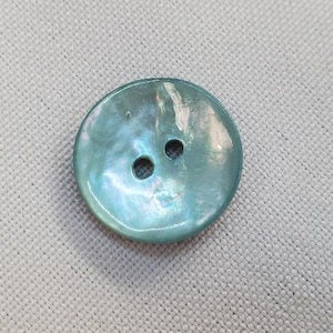 Pode incluir: Botão ovalado perolado com dois furos. O botão tem um brilho azul claro e iridescente, dando-lhe uma aparência sutil e brilhante. Está sobre um fundo de tecido cinza claro.