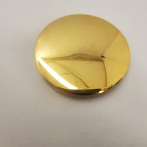 Op de afbeelding: Een glanzend, goudkleurig, cirkelvormig object met een licht gewelfd oppervlak. Het object reflecteert licht, waardoor highlights en schaduwen ontstaan. Het oppervlak heeft enkele onvolkomenheden. Het kan een decoratief element of onderdeel van een groter object zijn.