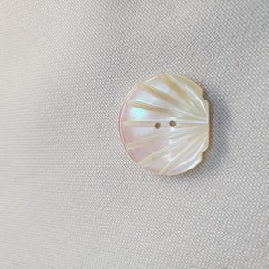 Könnte beinhalten: Perlmuttknopf in Halbkreisform mit Fächerdesign. Er hat zwei kleine Löcher zum Annähen und einen dezenten schillernden Glanz. Der Knopf ist hellrosa und cremefarben.