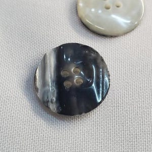 Puede incluir: Dos botones redondos sobre un fondo de tela blanca. Un botón es blanco nacarado y el otro es veteado en blanco y negro. Cada botón tiene cuatro agujeros para coser.