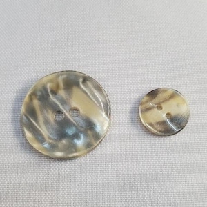Peut inclure: Deux boutons ronds avec un motif marbré crème et beige. Un bouton est plus grand, d'environ 3,8 cm de diamètre, et l'autre est plus petit, environ 1,9 cm de diamètre. Les deux ont deux trous.