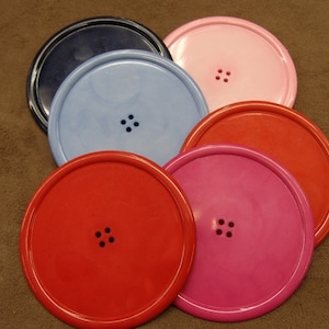 May include: Six round coasters with a button design. The coasters are in various colours, including blue, pink, red, and black.