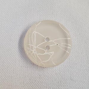 Puede incluir: Un botón circular semitranslúcido con una base de color beige claro y un patrón blanco distintivo en forma de telaraña sobre su superficie. El botón tiene dos orificios centrados para su fijación y un borde circular liso y ligeramente hundido.
