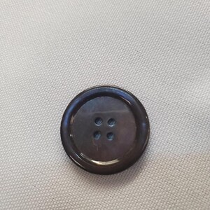 Peut inclure: Un bouton rond marron foncé avec quatre trous, posé sur un tissu beige clair. Le bouton a une finition brillante et un bord légèrement surélevé, suggérant un matériau durable.