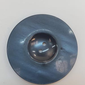 Peut inclure: Un bouton circulaire gris foncé avec une finition lisse et légèrement irisée. Le bouton présente une zone centrale en creux contenant deux petits trous alignés verticalement pour la fixation, entourée d'un large bord plat et poli.