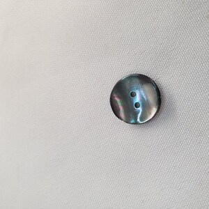 Op de afbeelding: Een ronde, iriserende knoop met twee gaatjes. De knoop heeft een donkere, metalen rand en een parelmoerachtige oppervlakte met tinten groen, blauw en paars. Het is geplaatst tegen een lichtgekleurde, getextureerde stoffen achtergrond.