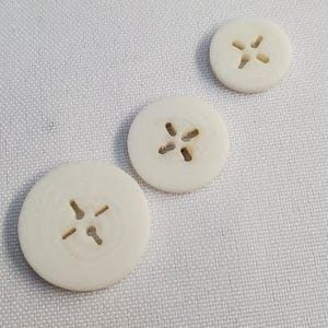 Può includere: Tre bottoni rotondi bianco sporco di diverse dimensioni sono disposti in diagonale. Ogni bottone presenta un disegno a forma di croce con quattro piccoli fori. I bottoni sono su uno sfondo di tessuto chiaro e strutturato.