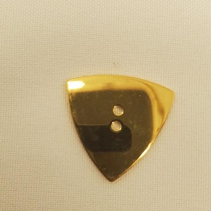 May include: A gold-colored guitar pick with a triangular shape and two small holes near the top. The pick is shiny and reflective, set against a textured, off-white background.