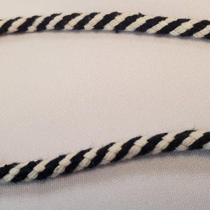 Puede incluir: Primer plano de un trozo de cuerda con un patrón en espiral blanco y negro. La cuerda está curvada, con los extremos fuera del encuadre. La cuerda está hecha de un material suave y tejido. El fondo es una superficie blanca lisa.