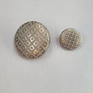 Pode incluir: Dois botões prateados com um padrão de losangos repetido. Um botão é grande e redondo, enquanto o outro é menor e oval. Os botões estão em um fundo branco.