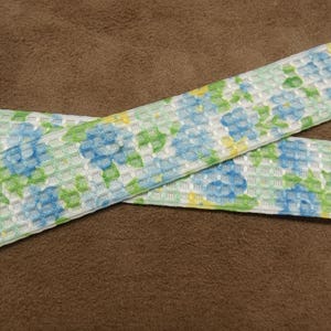Könnte beinhalten: Weißes Band mit einem blauen und grünen Blumenmuster. Das Band hat eine strukturierte Oberfläche und ist mit kleinen Quadraten gewebt.