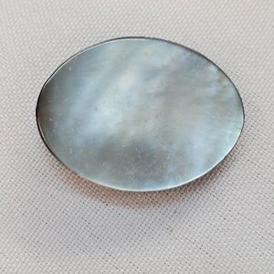 Op de afbeelding: Ovale, iriserende parelmoeren broche met een zilverkleurige metalen rand. De broche heeft een glad, gepolijst oppervlak dat licht reflecteert, waardoor een glinsterend effect ontstaat. Een klassiek accessoire.