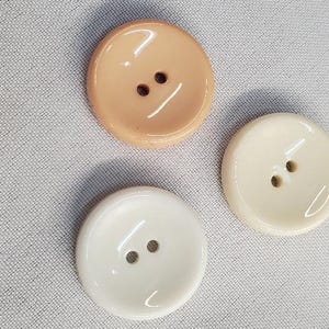 Puede incluir: Tres botones redondos en tonos beige y blanco. Cada botón tiene dos agujeros para coser. Los botones son brillantes y parecen estar hechos de un material liso. Los botones están sobre una tela gris claro.