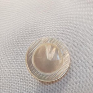 Pode incluir: Um botão redondo e iridescente com um círculo central ligeiramente elevado. A borda externa apresenta um padrão gravado diagonalmente. O botão é feito de um material translúcido, provavelmente concha ou similar.