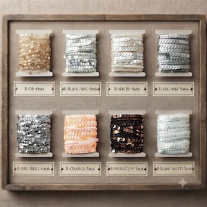 Op de afbeelding: Een houten displaybord met acht spoelen paillettenband in diverse kleuren zoals goud, zilver, wit, zwart en oranje. Elke spoel is voorzien van tekstlabels zoals R-OR-4MM, R-BLANC/ARG-5MM, R-NACRE-5MM, R-ARG-MAT-5MM, R-ARG-BRILL-6MM, R-ORANGE-5MM, R-NOIR/CUIV-5MM en R-BLANC-MULTI-5MM.