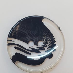 Puede incluir: Un botón redondo de dos agujeros con una base transparente y clara que presenta un patrón negro abstracto y arremolinado, creando un efecto fluido y jaspeado en la superficie. El botón tiene un acabado liso, brillante y un borde redondeado.