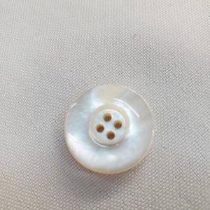 Peut inclure: Un bouton rond et nacré avec quatre petits trous bruns au centre. Le bouton a une finition brillante et est posé sur un fond de tissu beige clair. Idéal pour les projets de couture.