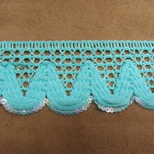 Peut inclure: Une garniture décorative turquoise avec un bord festonné. La garniture présente un motif de filet tissé au-dessus d'une série de motifs en forme de vagues. Le bord inférieur est orné de petites paillettes irisées.