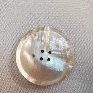Peut inclure: Gros bouton rond avec une finition nacrée. Le bouton a un éclat légèrement irisé et quatre petits trous près du centre pour la couture. Le bouton est fait d'une matière naturelle, peut-être une coquille.