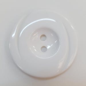 Può includere: Un bottone rotondo bianco a due fori con una finitura liscia e lucida. Il bottone presenta un'area centrale incavata contenente i due fori, circondata da un bordo esterno leggermente rialzato con un sottile motivo a onda inciso.