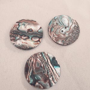 Peut inclure: Trois boutons ronds et irisés avec deux trous chacun. Les boutons ont un aspect naturel et texturé avec des nuances de marron, de sarcelle et de crème. Les boutons sont en coquille d'ormeau.