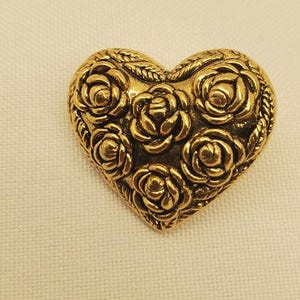 May include: A gold-coloured heart-shaped brooch with a floral rose design. The brooch is made of metal and features a detailed pattern of roses across its surface. The heart shape is outlined with a rope-like border.