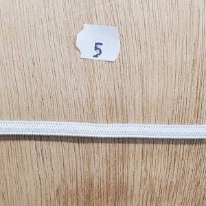 K&ouml;nnte beinhalten: Ein einzelnes, d&uuml;nnes, wei&szlig;es Gummiband, das horizontal ausgelegt ist. &Uuml;ber dem Gummiband befindet sich ein kleines, wei&szlig;es, rechteckiges Papieretikett mit der in schwarzer Tinte aufgedruckten Zahl 5, mittig im oberen Bereich platziert.