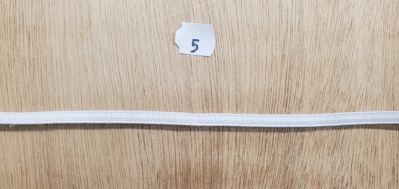 K&ouml;nnte beinhalten: Ein einzelnes, d&uuml;nnes, wei&szlig;es Gummiband, das horizontal ausgelegt ist. &Uuml;ber dem Gummiband befindet sich ein kleines, wei&szlig;es, rechteckiges Papieretikett mit der in schwarzer Tinte aufgedruckten Zahl 5, mittig im oberen Bereich platziert.