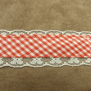 May include: A red and white gingham ribbon with white lace trim. The ribbon is on a brown background.