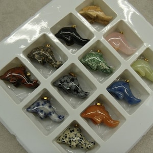 May include: A white tray with 12 dolphin-shaped pendants made of various types of stone. The pendants are arranged in a grid pattern and each one has a gold loop for hanging.