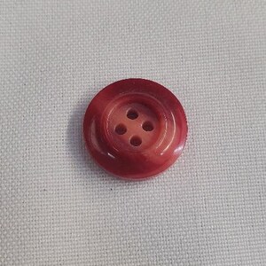 May include: A round, deep red button featuring a smooth, glossy finish and a recessed center. The button has four small, evenly spaced holes arranged in a square pattern in the middle, designed for sewing onto garments or other fabric items.