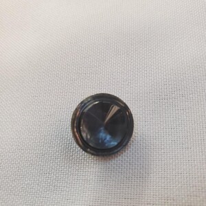 Puede incluir: Un botón gris oscuro con un diseño circular. El botón tiene un borde circular en relieve y un centro facetado y reflectante. El botón está sobre un fondo de tela blanca.