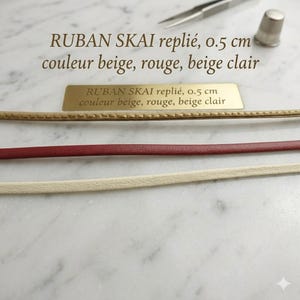 May include: Three strips of folded faux leather ribbon in beige, red, and light beige, each measuring 0.5 cm in width. The text RUBAN SKAI repli&eacute;, 0.5 cm couleur beige, rouge, beige clair is printed on the image and on a gold metal plaque.