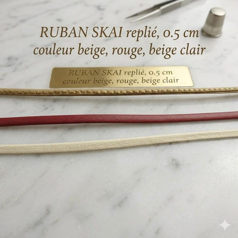 May include: Three strips of folded faux leather ribbon in beige, red, and light beige, each measuring 0.5 cm in width. The text RUBAN SKAI repli&eacute;, 0.5 cm couleur beige, rouge, beige clair is printed on the image and on a gold metal plaque.