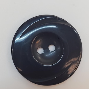 Peut inclure: Un bouton noir brillant et rond doté d'un centre lisse légèrement concave avec deux trous centrés pour la fixation. Le bouton présente une finition polie qui reflète la lumière, créant des reflets subtils sur sa surface circulaire incurvée.