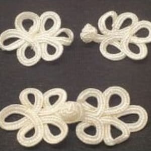 Peut inclure: Trois fermetures à grenouille blanches en forme de fleur. Deux sont séparées et une est connectée. Les fermetures sont faites d'un matériau tissé et sont présentées sur un fond sombre. Ces fermetures sont souvent utilisées à des fins décoratives sur les vêtements.
