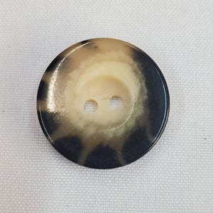 Puede incluir: Un botón ovalado grande con dos agujeros. El botón tiene un centro de color crema y un anillo exterior moteado marrón oscuro. El botón está sobre un fondo blanco texturizado.