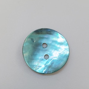 Pode incluir: Um botão redondo de dois furos com uma superfície iridescente e cintilante em tons profundos de azul-petróleo, azul e verde. O material exibe um brilho perolado natural com padrões em espiral por toda a face circular do fecho.